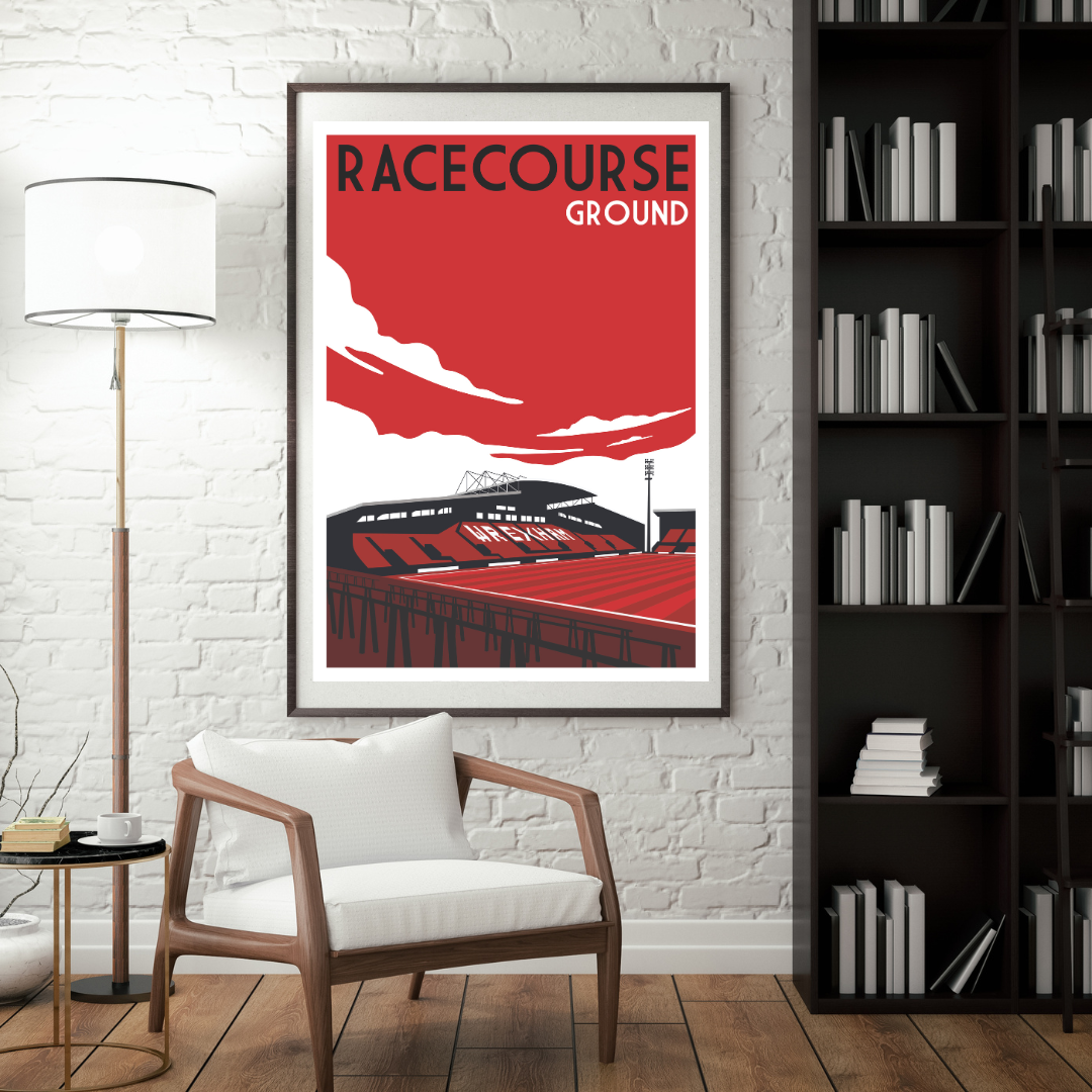 a framed poster of a stadium with a red and white color scheme, hanging on a white brick wall. In front of the poster, there is a white chair, and a bookshelf filled with books is visible to the right of the poster.