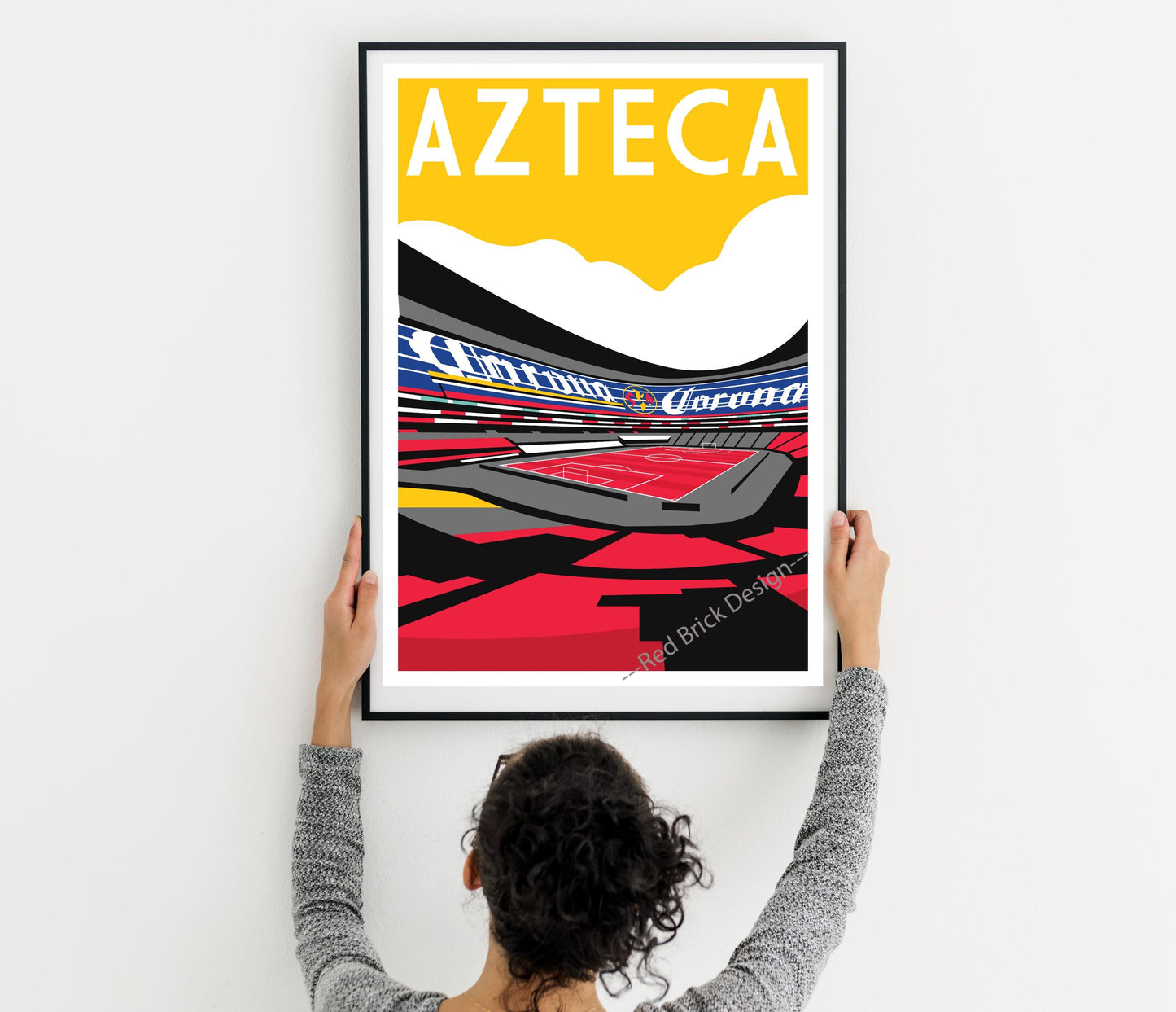 A person is holding up a framed poster of an Aztec stadium, which features a colorful and stylized illustration of the stadium.