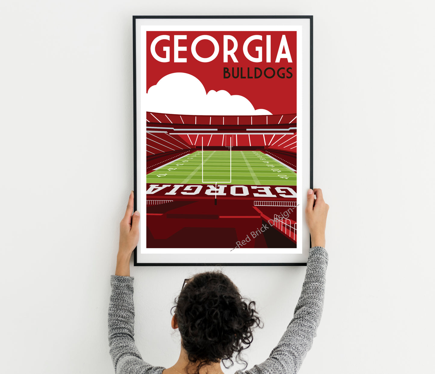 A person is hanging a framed poster of a football stadium in Georgia, with the text "GEORGIA BULLDOGS" prominently displayed.