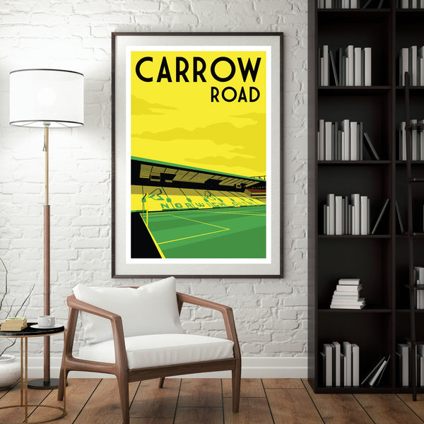 a framed poster of a soccer stadium on a wall, with a white chair and a bookshelf in the foreground.