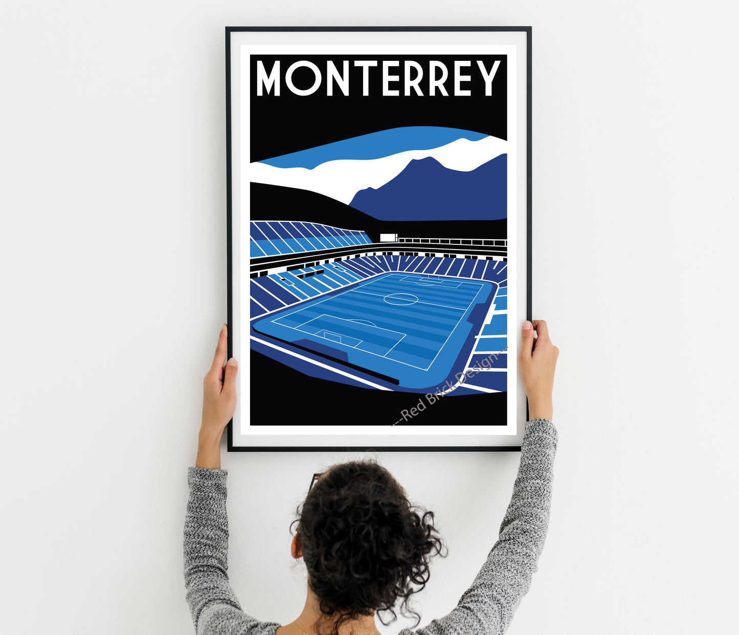 A person is holding up a framed poster of a soccer stadium, which features a mountain landscape in the background.