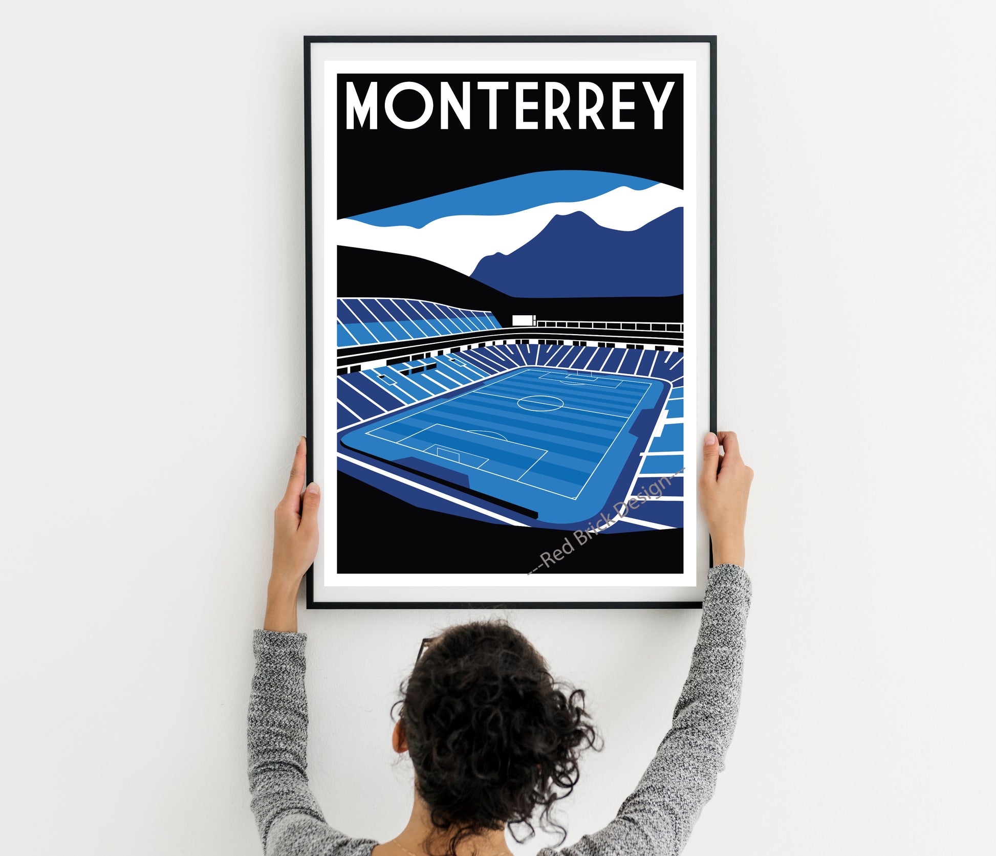 A person is holding up a framed poster of a soccer stadium, which features a mountain landscape in the background.
