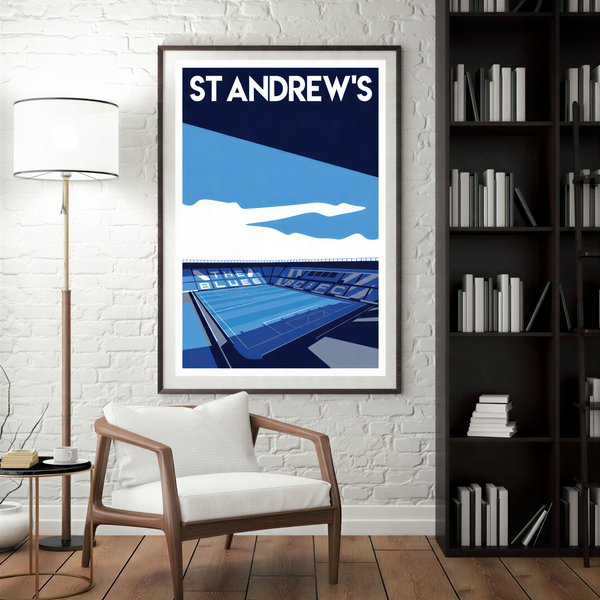a framed poster of St Andrews' stadium, with a white chair in the foreground and a bookshelf filled with books in the background.