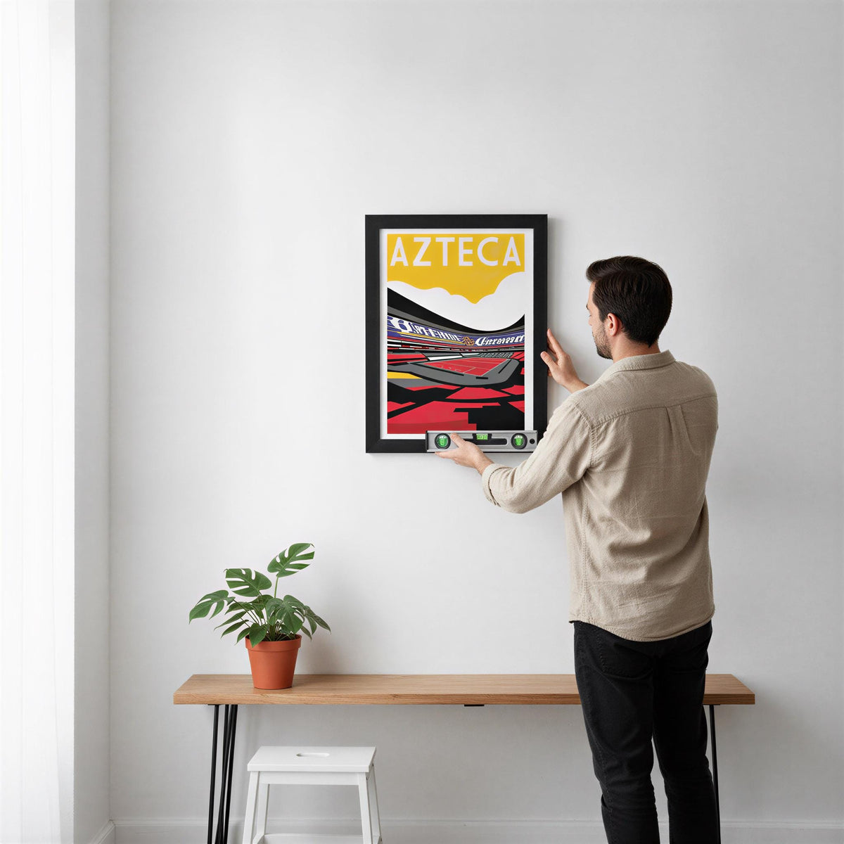 A man is hanging a framed poster of Azteca, a region in Latin America, on a white wall.