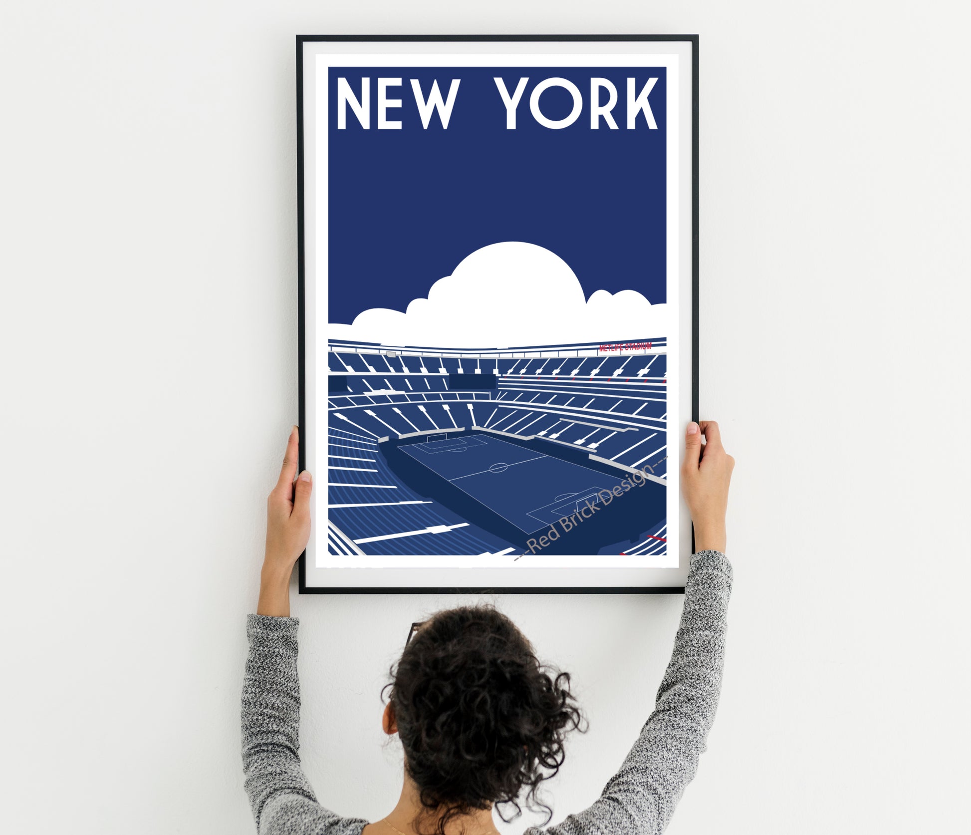 A person is hanging a framed poster of a stadium on a wall.