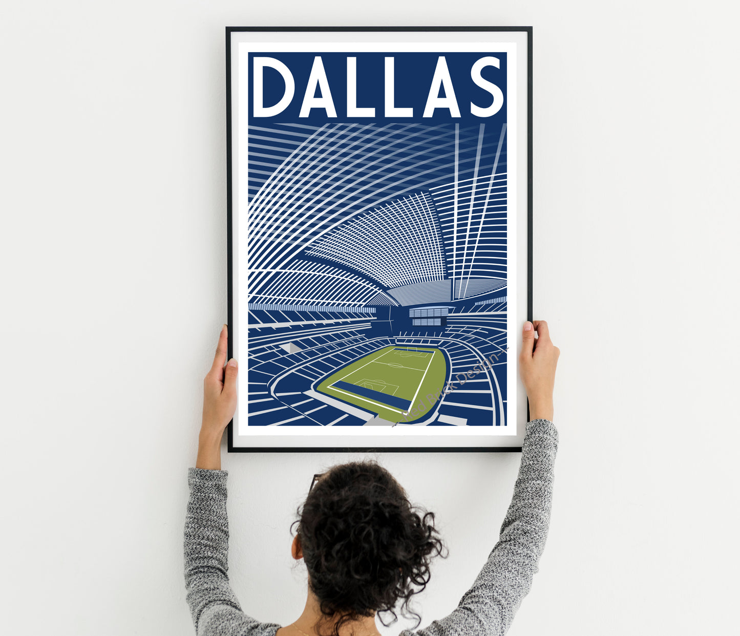 A person is hanging a framed poster of a stadium with the word "DALLAS" written on it.