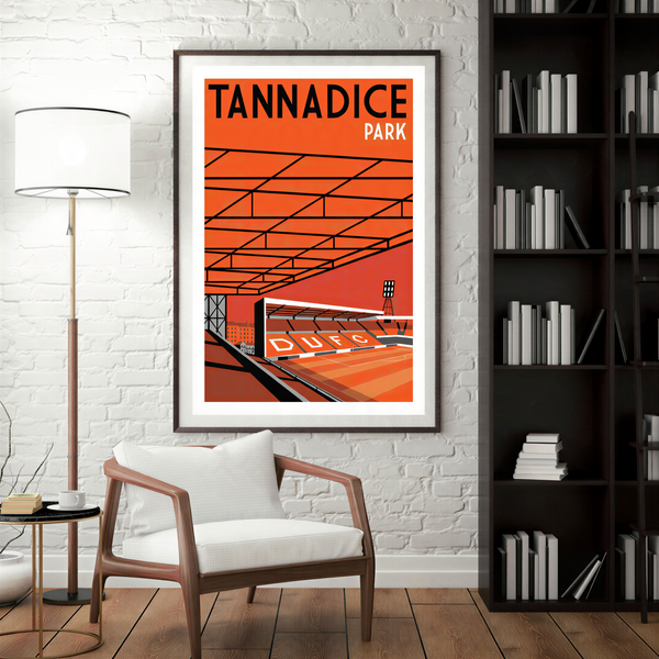 a framed poster of a stadium with the text "TANNADICE PARK" on it, hanging on a white brick wall. In the foreground, there is a white chair, and a bookshelf filled with books can be seen in the background.