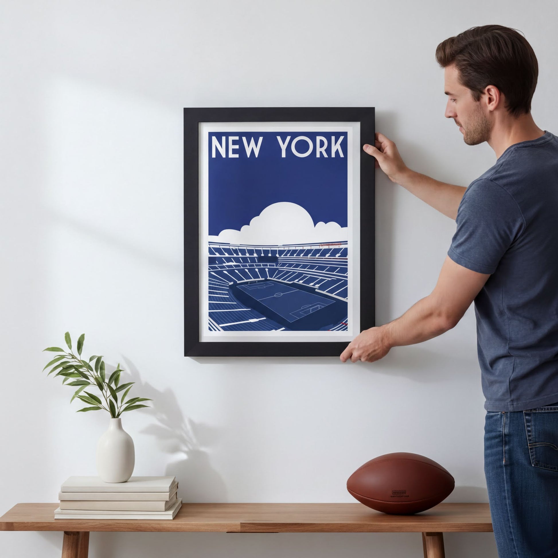 A man is hanging a framed poster of a stadium on a wall, with a potted plant, books, and a football nearby.