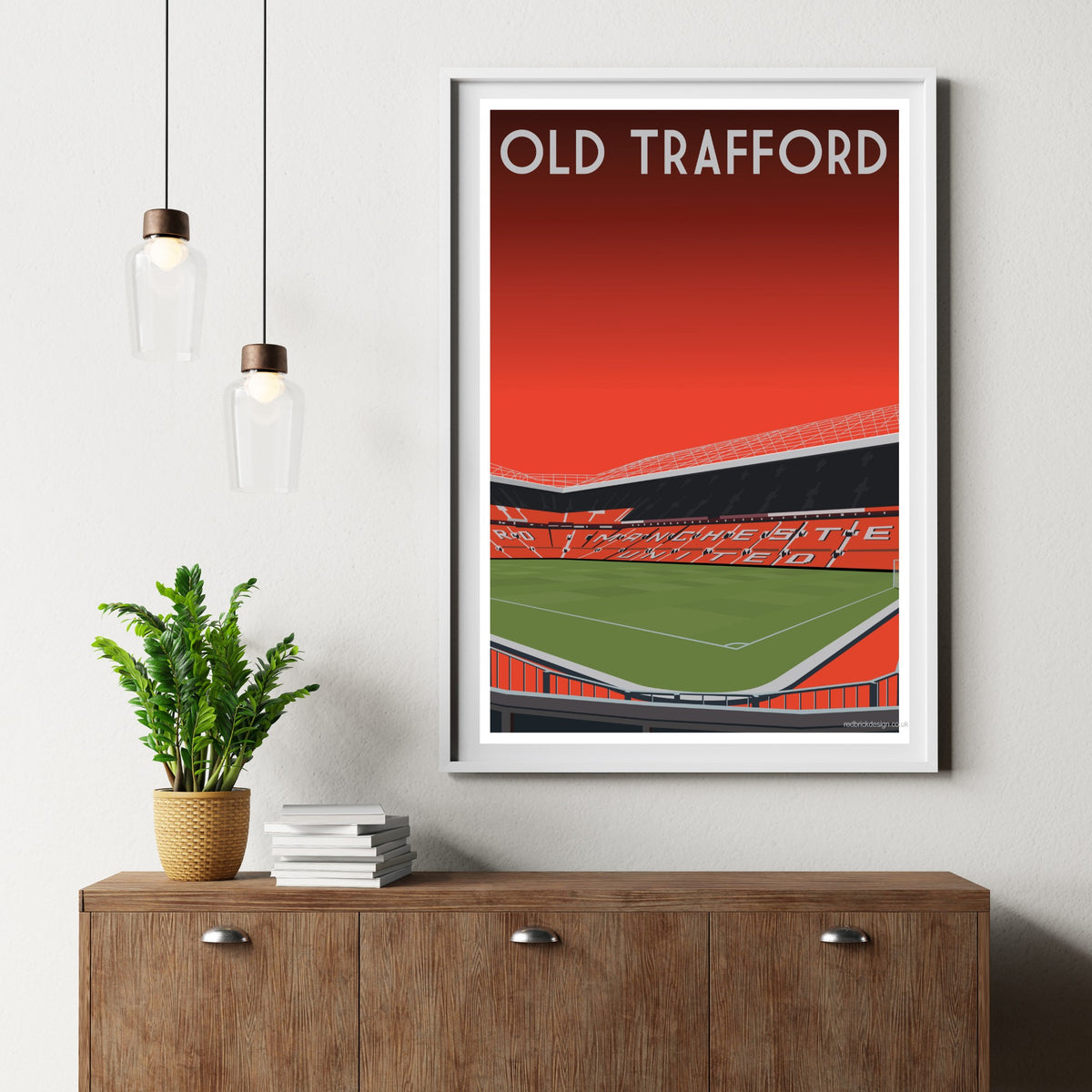 a framed poster of a football stadium, specifically the Old Trafford stadium, hanging on a wall above a wooden cabinet.