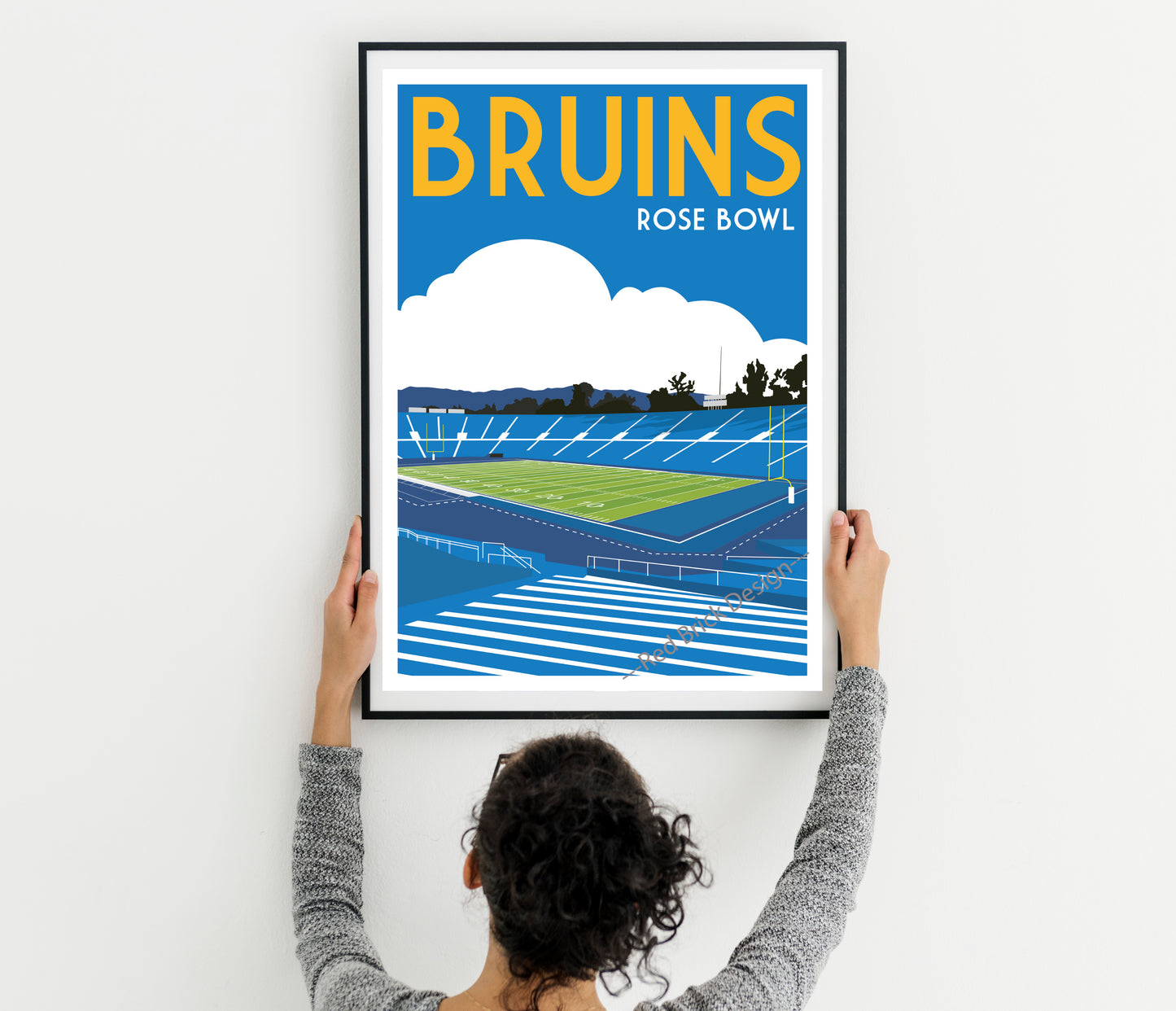 A person is hanging a framed poster of a sports stadium, specifically the Bruins Rose Bowl, on a wall.