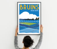 A person is hanging a framed poster of a sports stadium, specifically the Bruins Rose Bowl, on a wall.