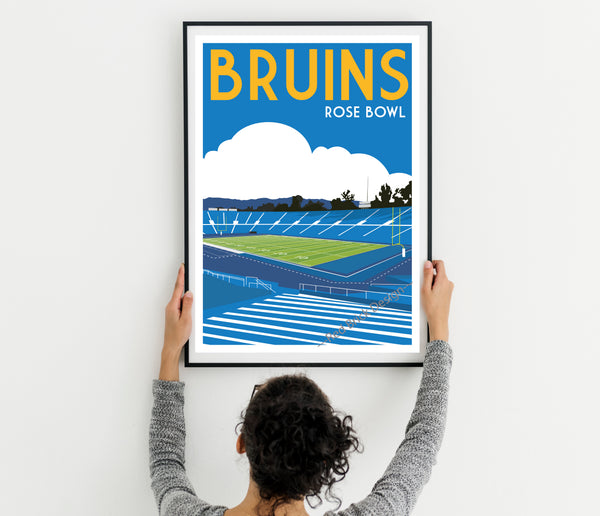A person is hanging a framed poster of a sports stadium, specifically the Bruins Rose Bowl, on a wall.
