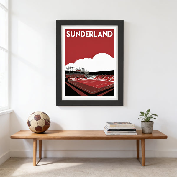 A framed poster of a stadium is hanging on a white wall above a wooden bench, with a soccer ball and some books placed on the bench.