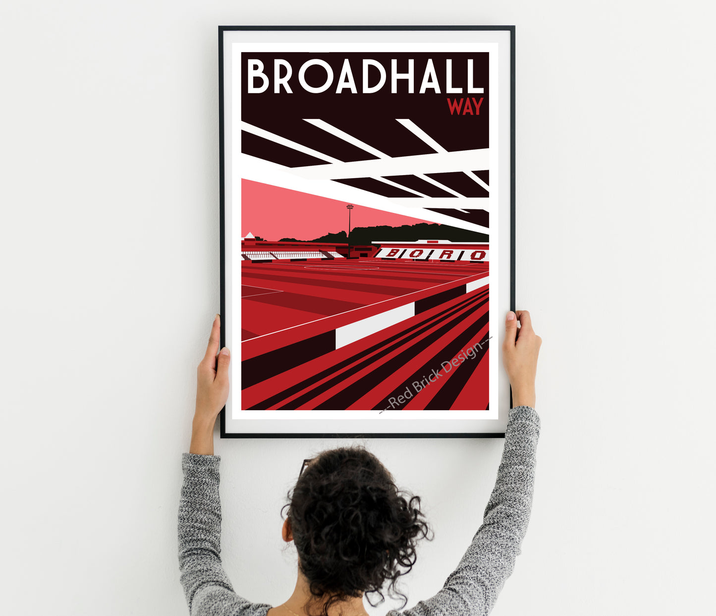 A person is holding up a framed poster of Broad Hall Way, which features a red and black design with a stadium and track.