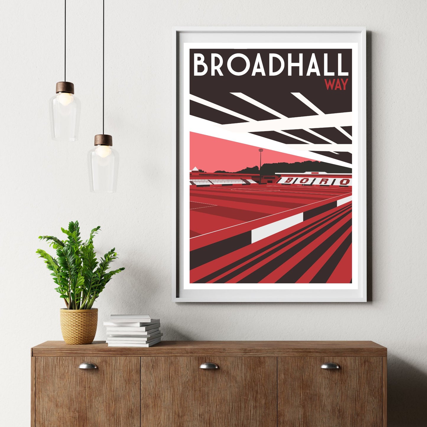 a framed poster of Broadhall Way, a football stadium, hanging on a wall above a wooden dresser with a stack of books and a potted plant.