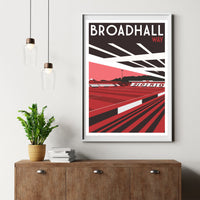 a framed poster of Broadhall Way, a football stadium, hanging on a wall above a wooden dresser with a stack of books and a potted plant.