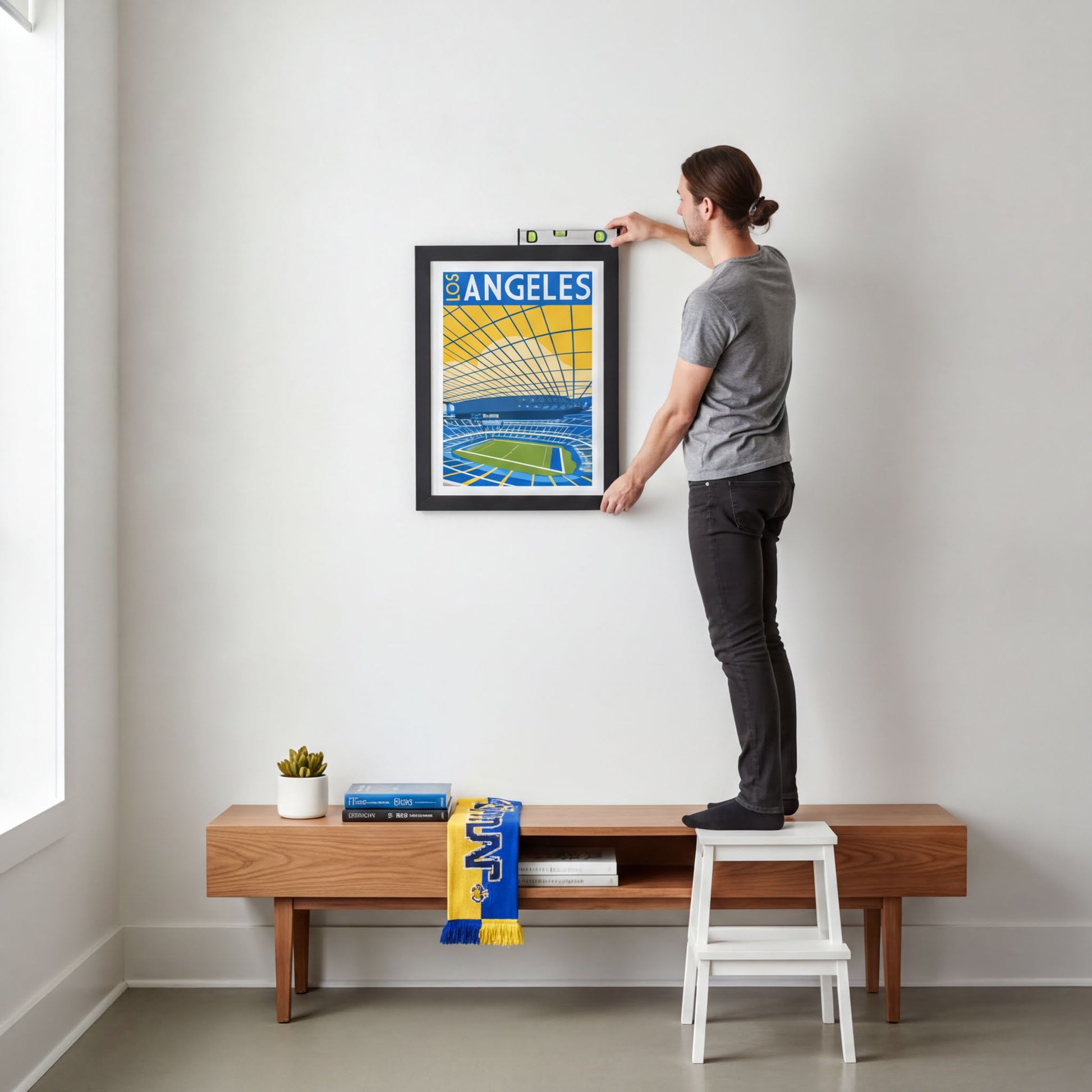 A person is standing on a wooden bench, measuring and hanging a framed poster of a stadium on a white wall.