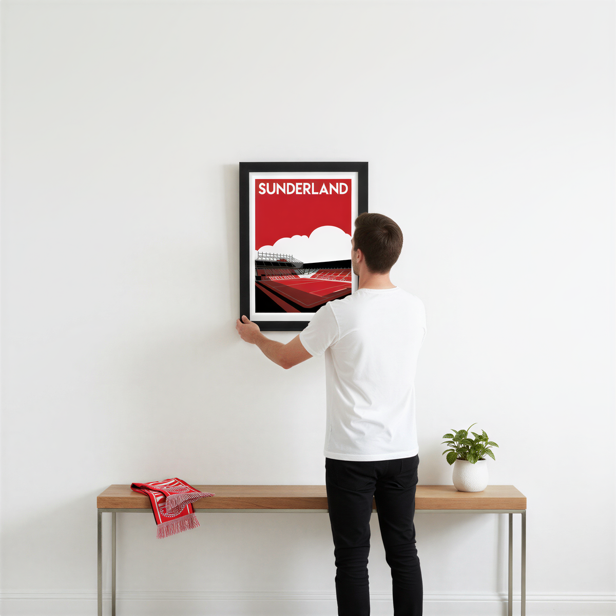 A man is hanging a framed poster of a stadium on a white wall.