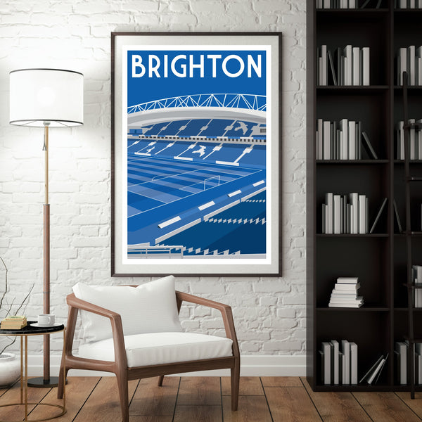a framed poster of a stadium with the word "BRIGHTON" written on it, hanging on a white brick wall. In front of the poster, there is a white chair, and a bookshelf filled with books can be seen in the background.