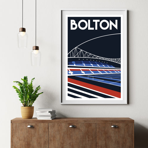 a framed poster of a stadium with a bridge in the background, hanging on a wall above a wooden dresser with a stack of books and a potted plant.