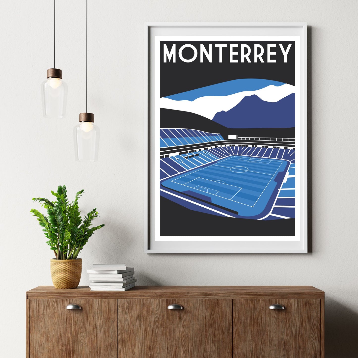 a framed poster of a soccer stadium, with a potted plant and a stack of books on a wooden dresser below it.
