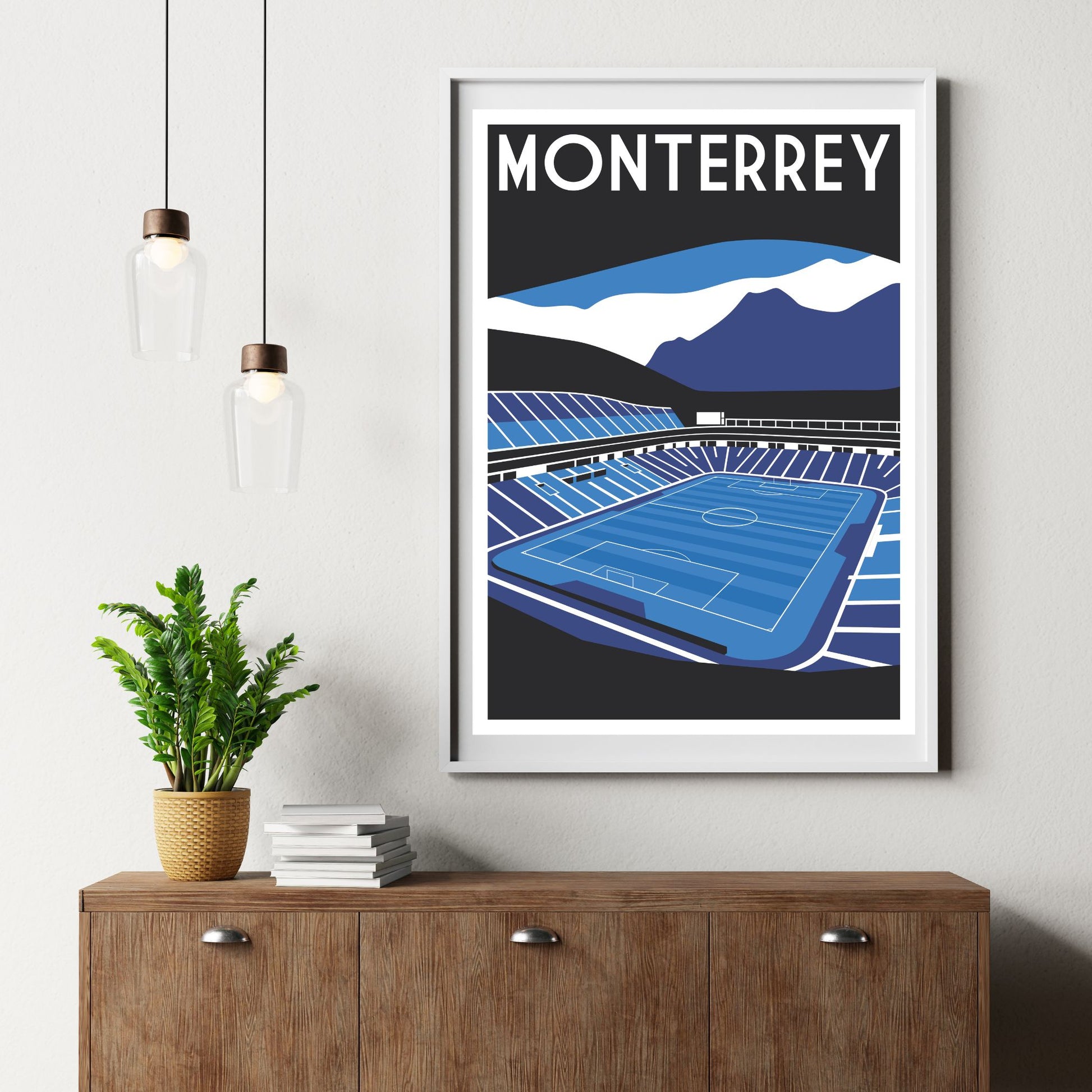 a framed poster of a soccer stadium, with a potted plant and a stack of books on a wooden dresser below it.