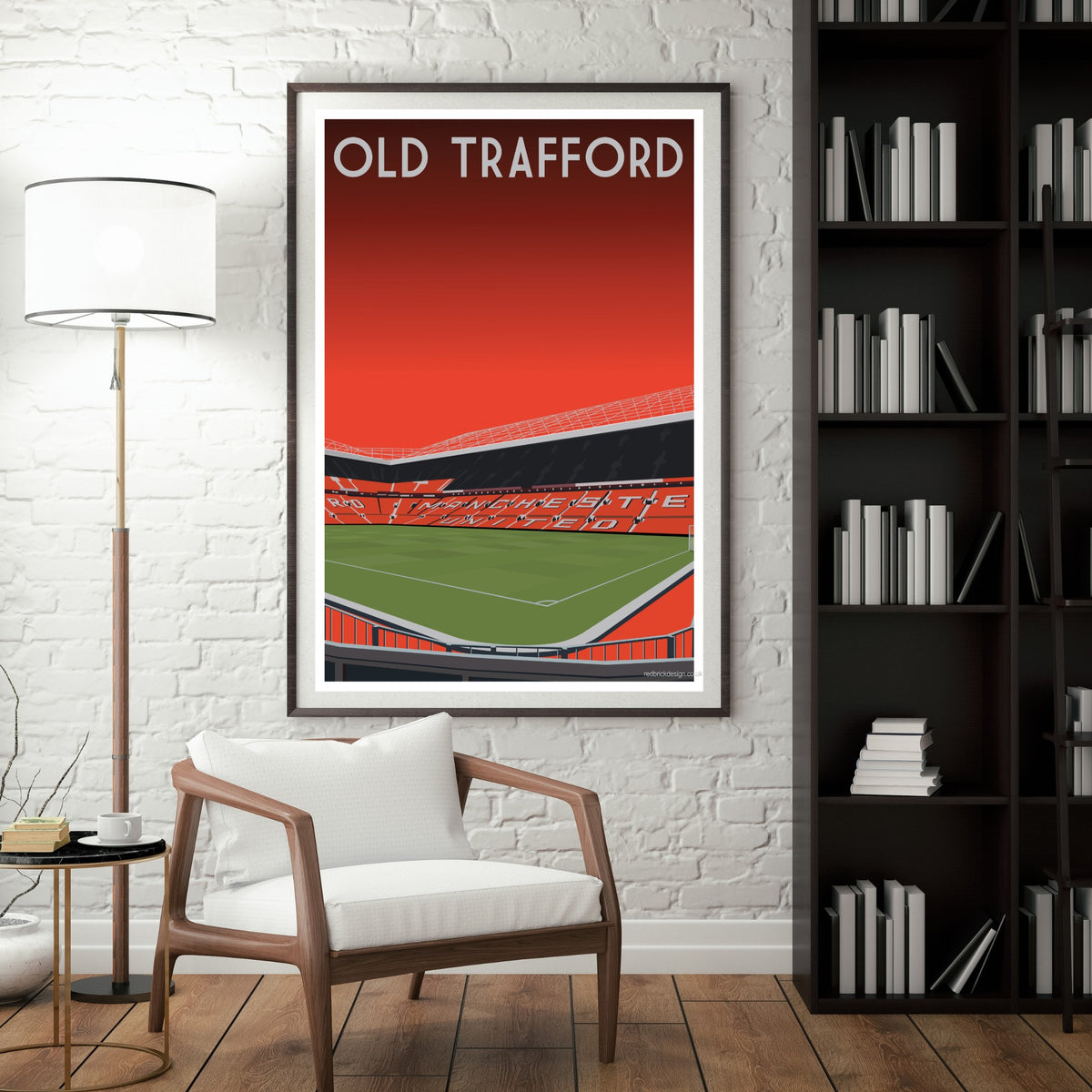 a framed poster of a football stadium, specifically the Old Trafford stadium, hanging on a wall in a room with a white chair and a bookshelf.