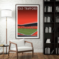 a framed poster of a football stadium, specifically the Old Trafford stadium, hanging on a wall in a room with a white chair and a bookshelf.
