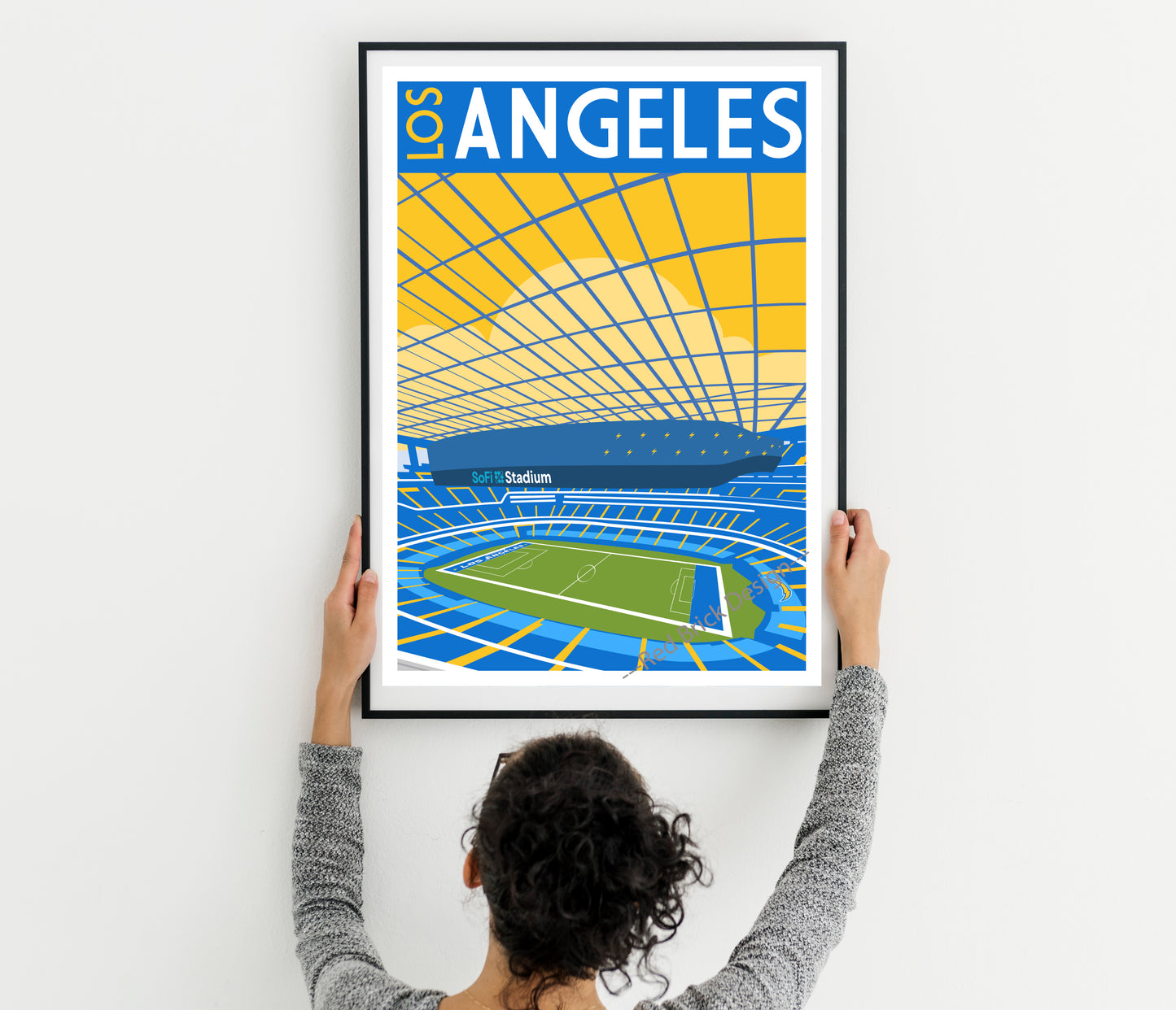 A person is hanging a framed poster of a stadium on a wall.