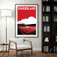 A framed poster of a stadium is hanging on a white brick wall, with a white chair in the foreground and a bookshelf filled with books to the right.