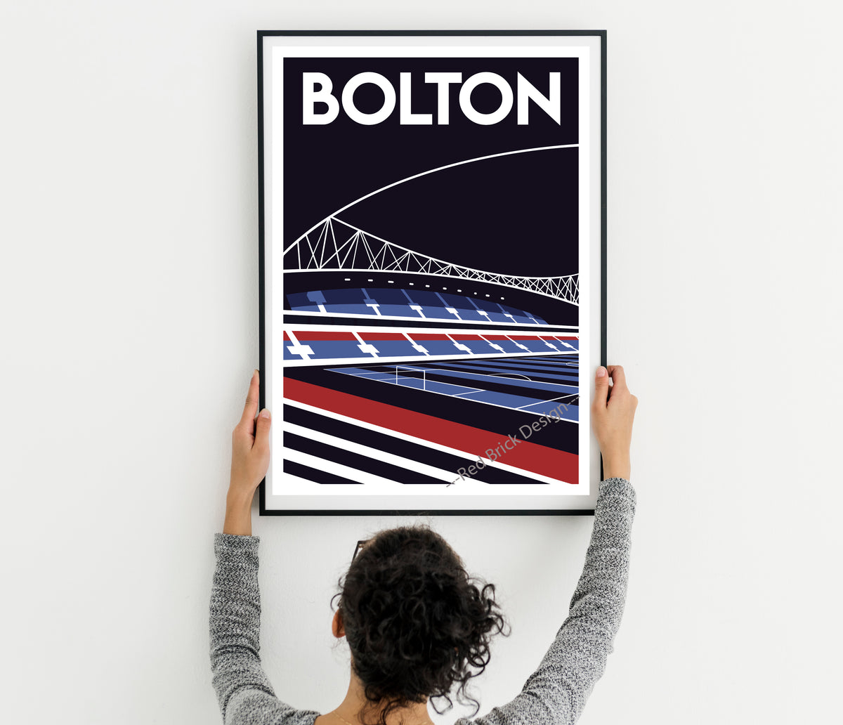 A person is hanging a framed poster of a stadium on a wall.