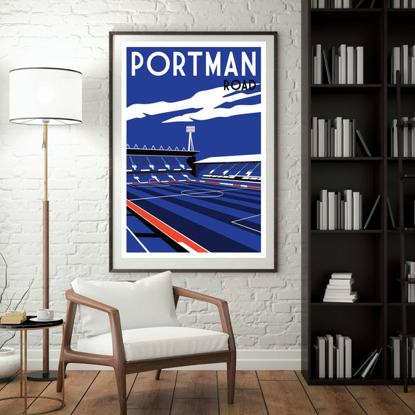 a framed poster of a stadium with a blue sky and clouds, hanging on a white brick wall. In front of the poster, there is a white chair and a bookshelf filled with books.