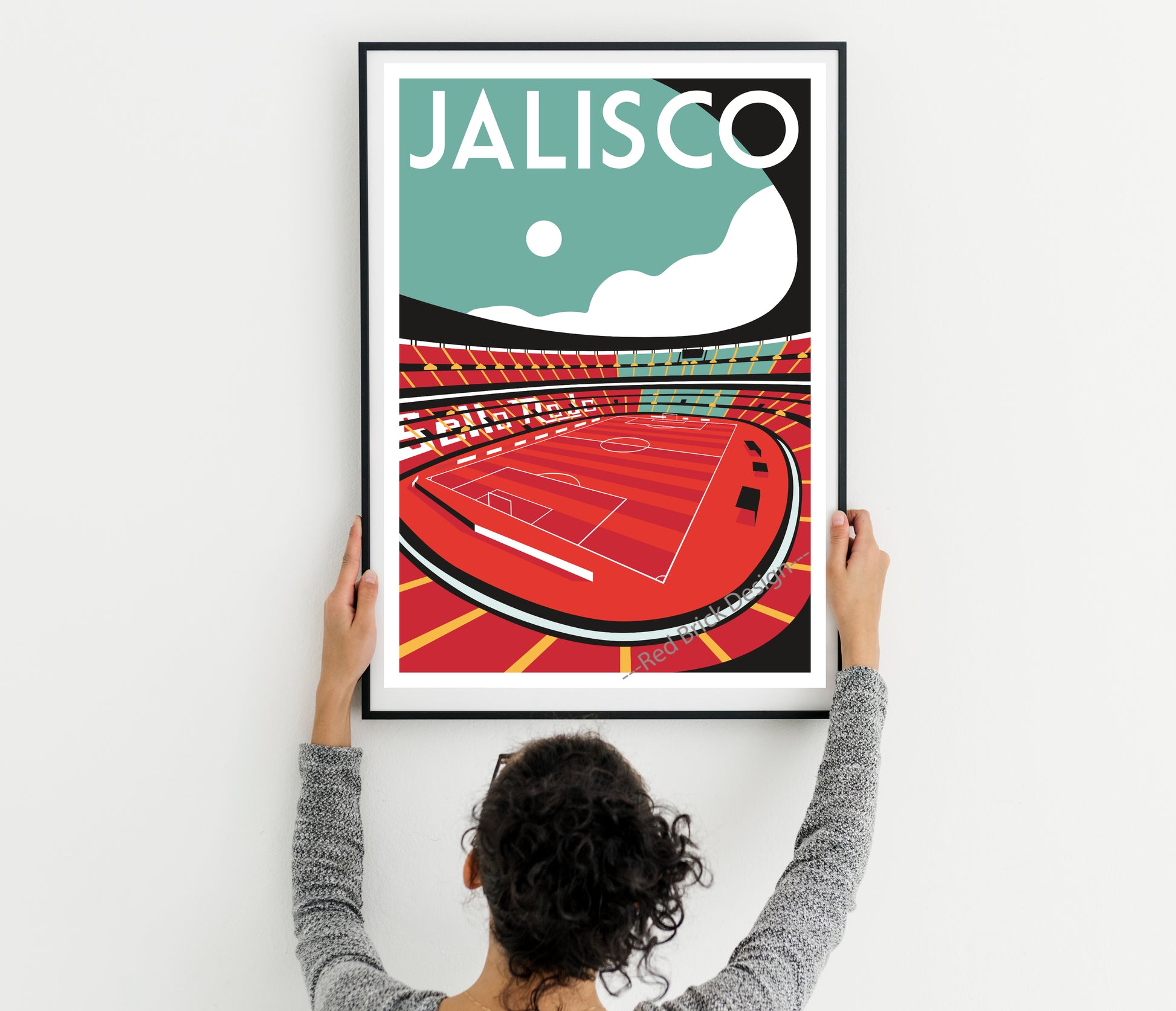 A person is hanging a framed poster of a soccer stadium on a wall.