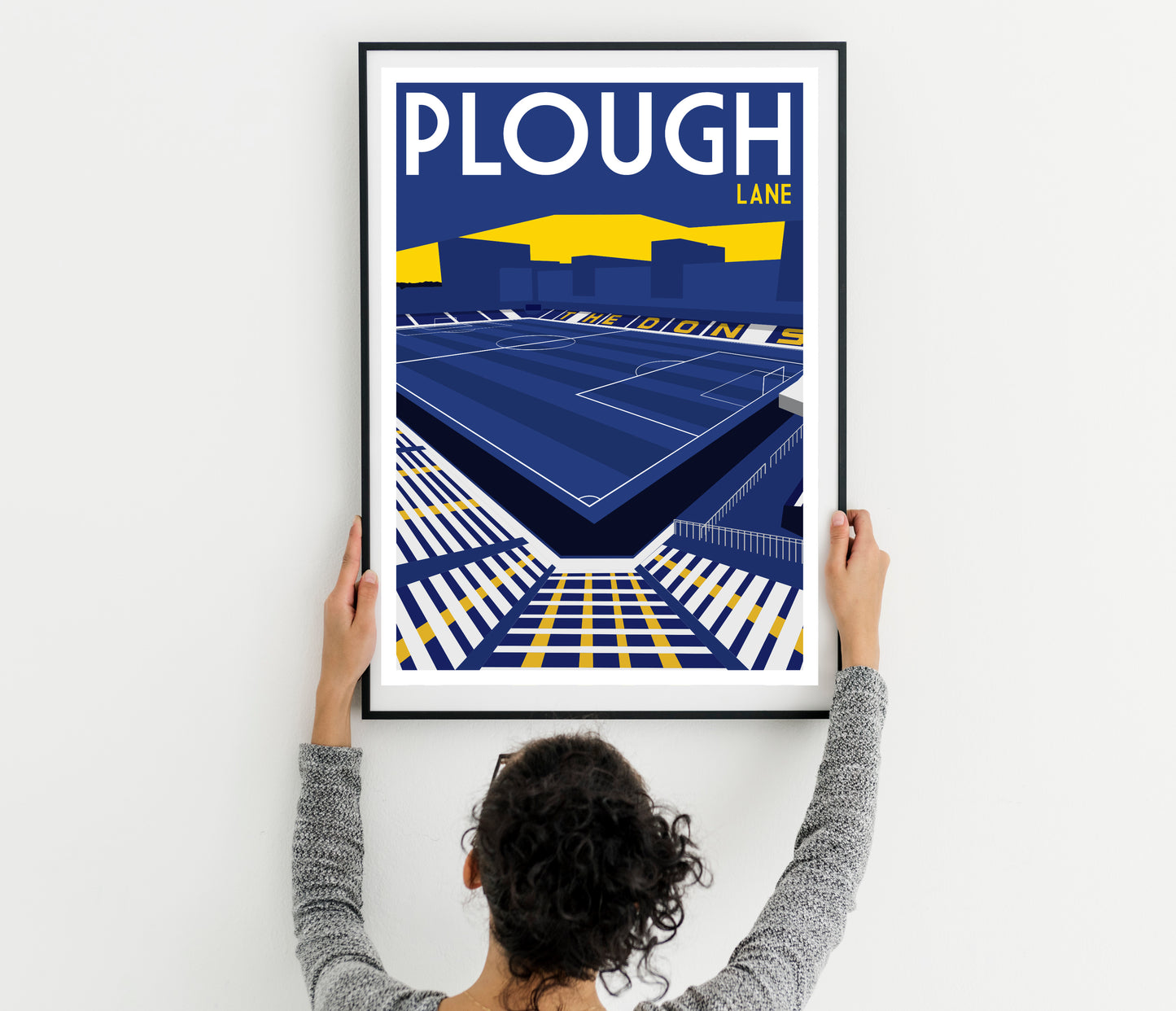 A person is hanging a framed poster of a stadium on a wall.