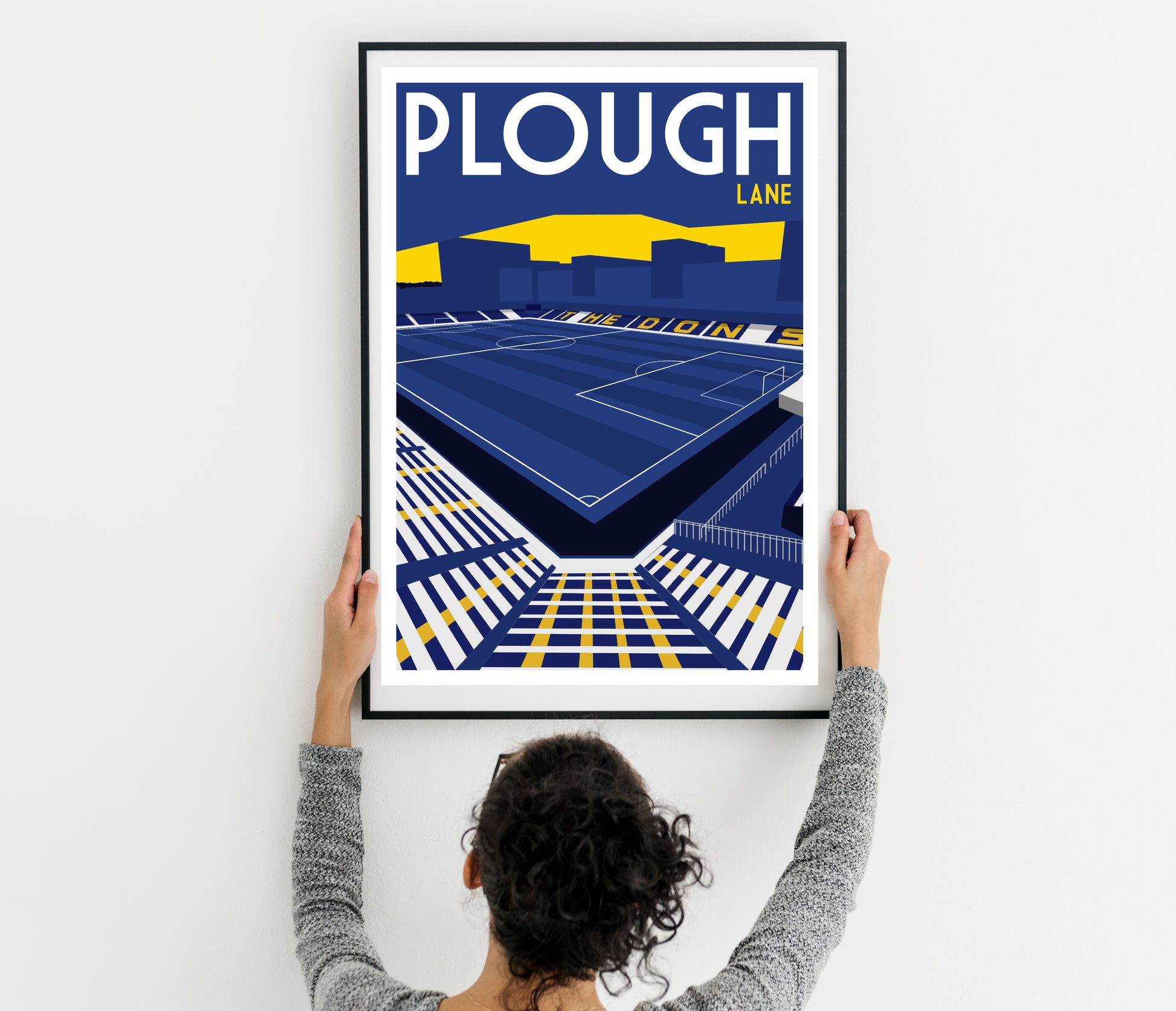A person is hanging a framed poster of a stadium on a wall.