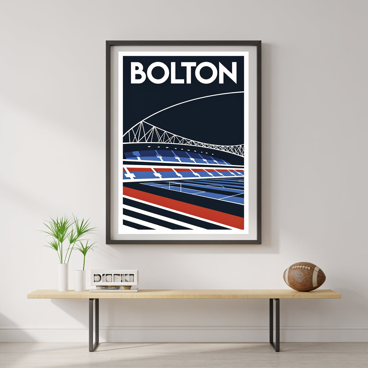 a framed poster of a stadium with a football field, hanging on a wall above a wooden table with a plant and a football.