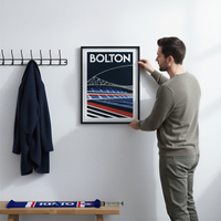 A man is hanging a framed poster of Bolton on a wall, with a coat rack and a bench visible in the background.