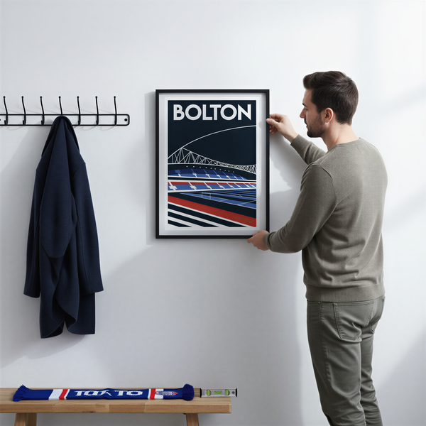A man is hanging a framed poster of Bolton on a wall, with a coat rack and a bench visible in the background.