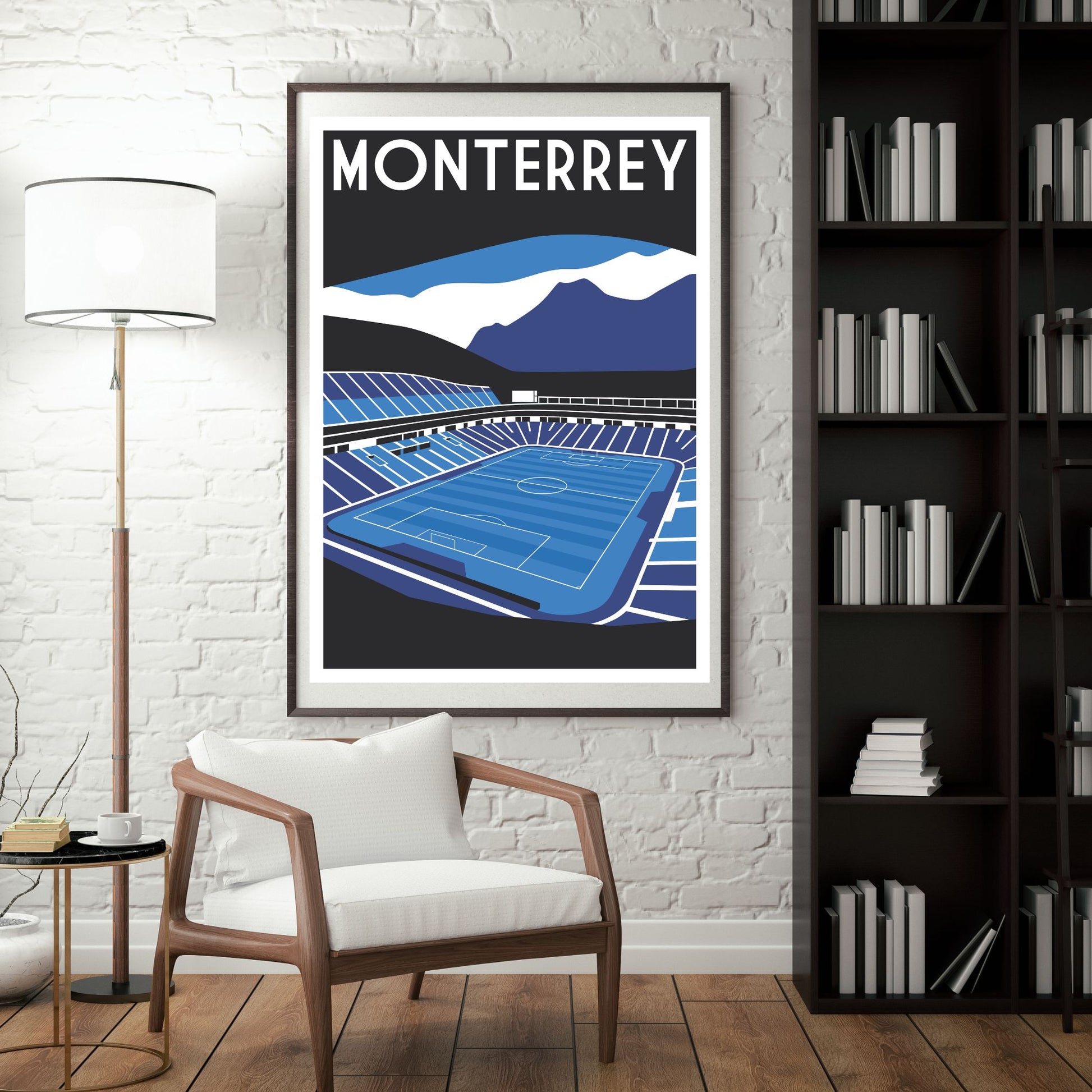 a framed poster of a soccer stadium, with mountains in the background, hanging on a wall next to a bookshelf.