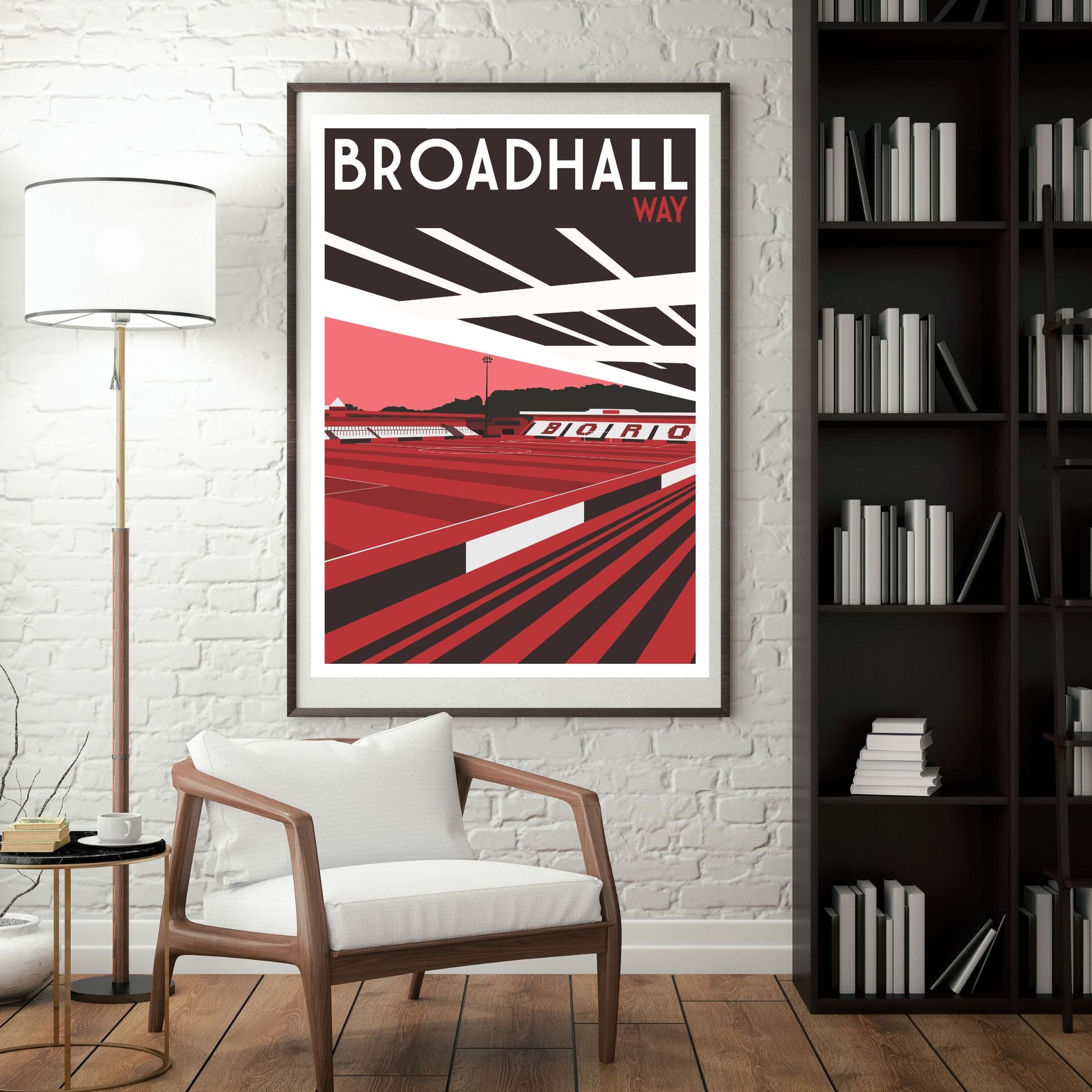 a framed poster of a stadium with the text "BROADHALL WAY" on it, hanging on a white brick wall. In front of the poster, there is a white chair, and a bookshelf filled with books can be seen in the background.